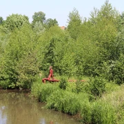 Skulptur "Der rote Mann" Eine rote Skulptur steht am Flussufer, umgeben von üppigem Grün, inmitten einer friedvollen Landschaft.