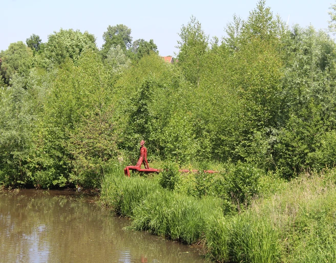 Skulptur "Der rote Mann" Eine rote Skulptur steht am Flussufer, umgeben von üppigem Grün, inmitten einer friedvollen Landschaft.