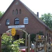 Haus der Vielfalt Backsteinhaus mit drei Fensterbögen und "Haus der Vielfalt"-Schild, umgeben von blühenden Pflanzen.