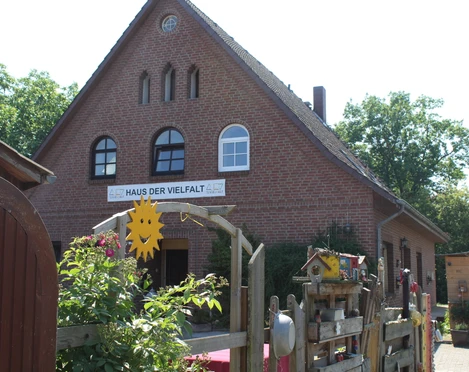 Haus der Vielfalt Backsteinhaus mit drei Fensterbögen und "Haus der Vielfalt"-Schild, umgeben von blühenden Pflanzen.