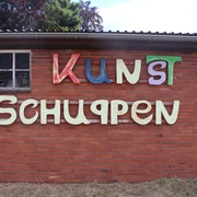 Kunstschuppen Asendorf Bunte Buchstaben auf rotem Backstein, Holzfenster links, Kunstschuppen-Schriftzug mittig.