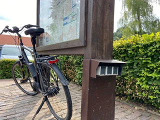 E-Bike-Ladestation Rathaus3.jpg Ein E-Bike lehnt an einer hölzernen Infotafel. Daneben ist eine Ladestation erkennbar.