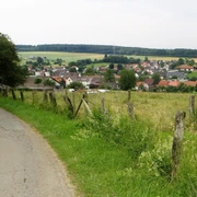 Brüder-Grimm-Weg Ländlicher Weg mit Blick auf ein Dorf im Hintergrund, umgeben von grünen Wiesen und bewaldeten Hügeln.