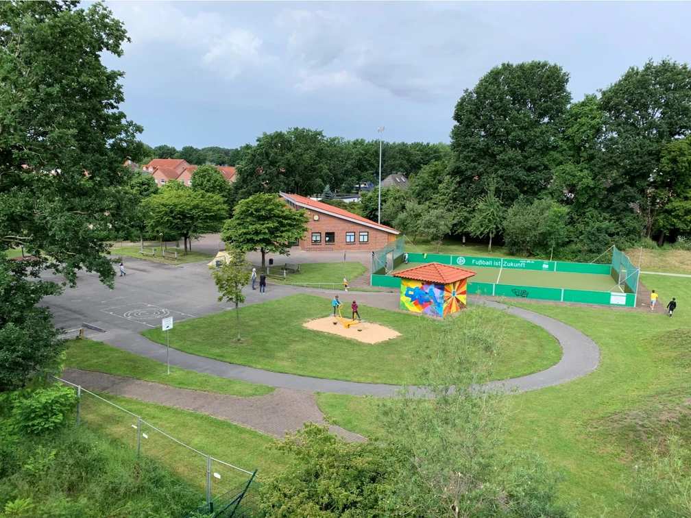 Außenansicht CASA Das Bild zeigt einen kleinen Spielplatz mit Sandkasten und bunter Hütte zwischen Bäumen, nahe einem roten Backsteingebäude und einem eingezäunten Sportplatz.