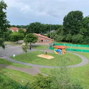 Außenansicht CASA Das Bild zeigt einen kleinen Spielplatz mit Sandkasten und bunter Hütte zwischen Bäumen, nahe einem roten Backsteingebäude und einem eingezäunten Sportplatz.