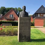 Skulptur von Adelbert Hotzen steht vor einem Ziegelhaus, umgeben von gepflegten Grünflächen.
