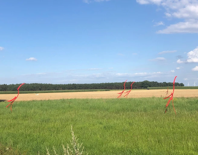 Skulptur Nestbeobachter Rote, abstrakte Stabfiguren aus Metall stehen auf einer grünen Wiese unter einem klaren blauen Himmel.