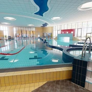 Ein modernes Schwimmbad bietet ein großes Becken mit klar abgegrenzten Schwimmbahnen und eine sanfte, angenehme Beleuchtung, die durch Fenster Tageslicht hereinlässt. Das Design ist einladend und barrierefrei gestaltet.