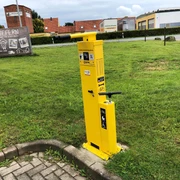 Lufttankstelle Hoya classic Gelbe Fahrrad-Luftstation am Gehweg, umgeben von Grünflächen und Backsteingebäuden im Hintergrund.