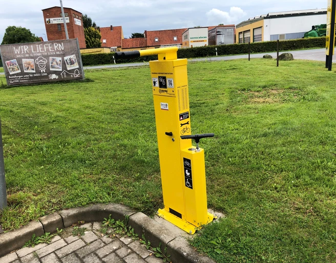 Lufttankstelle Hoya classic Gelbe Fahrrad-Luftstation am Gehweg, umgeben von Grünflächen und Backsteingebäuden im Hintergrund.