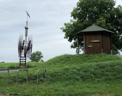 Skulptur Marschbodenglück - Schibboleth Skulptur mit kunstvoller Treppe und Windspiel, neben hölzernem Pavillon auf grünem Hügel.