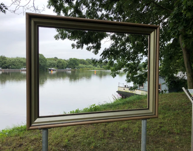 Weserblick 2021 Ein goldener Bilderrahmen steht am Weserufer, mit Blick auf den Fluss und die bewaldeten Uferbereiche.