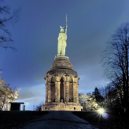 Beleuchtetes Hermannsdenkmal bei Nacht, umgeben von Bäumen, unter einem klaren Nachthimmel.