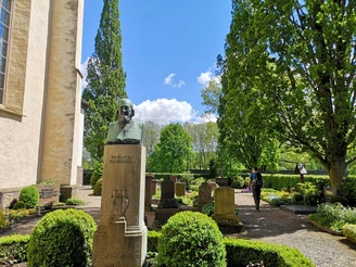 Friedhof Corvey mit dem Grab des Heinrich Hoffmann von Fallersleben Bronzebüste von Heinrich Hoffmann von Fallersleben auf einem Friedhof, umgeben von grünen Bäumen.
