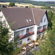 Luftaufnahme.jpg Luftbild eines weißen Wohnhauses mit braunem Dach und blühenden Blumenkästen vor Waldkulisse.