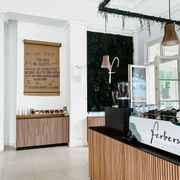 Innenansicht des Cafés mit der Theke rechts, auf der Gläser, Tassen und eine Kaffeemaschine stehen, und kleinen Regal geradeaus, über dem ein Poster mit handschriftlichem Text hängt. Von der Decke hängen zwei Lampen. Die Wände sind hoch.