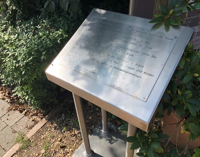 Synagoge Hoya Gedenktafel für die Synagoge Hoya, umrahmt von Pflanzen, auf einem schattigen Platz.