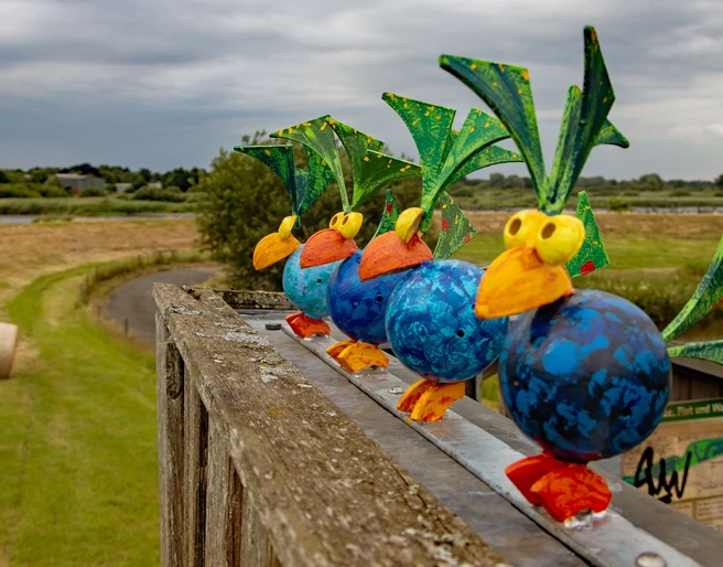 Bunte Vogelschar am Deich Eine Reihe bunter Vogel-Skulpturen auf einem Geländer, mit einer grünen Landschaft im Hintergrund.