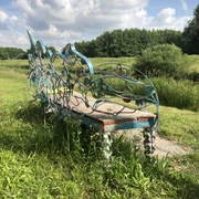 Lieblingsort Heeren-Wehren-Brücke Eine kunstvoll verzierte Bank steht auf einer Wiese, umgeben von üppigem Grün und einem blauen Himmel im Hintergrund. Die geschwungenen Metallverzierungen sind in bunten Farben gehalten und verleihen der Szenerie eine künstlerische Note.