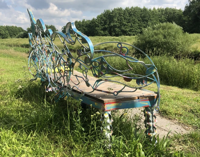 Lieblingsort Heeren-Wehren-Brücke Eine kunstvoll verzierte Bank steht auf einer Wiese, umgeben von üppigem Grün und einem blauen Himmel im Hintergrund. Die geschwungenen Metallverzierungen sind in bunten Farben gehalten und verleihen der Szenerie eine künstlerische Note.