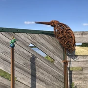 Reiher und Frosch  Das Bild zeigt eine stilisierte Reiherfigur aus rostfarbenen Stahll, die auf einem Holzzaun sitzt. Daneben ist ein kleiner, bunt gestalteter Frosch dargestellt, der ebenfalls auf einer Metallstange befestigt ist. Der Hintergrund besteht aus blauem Himmel und grüner Landschaft.