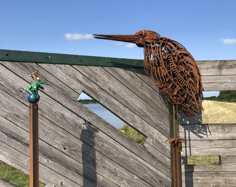 Reiher und Frosch  Das Bild zeigt eine stilisierte Reiherfigur aus rostfarbenen Stahll, die auf einem Holzzaun sitzt. Daneben ist ein kleiner, bunt gestalteter Frosch dargestellt, der ebenfalls auf einer Metallstange befestigt ist. Der Hintergrund besteht aus blauem Himmel und grüner Landschaft.