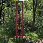 Lieblingsort Unterster Weg  Schmale rote Metallskulptur mit abstrakten Formen, vor dichtem grünem Wald aufgestellt.