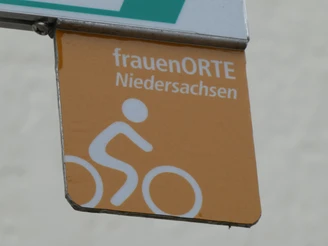 Logo Radweg frauenORTE Braunes Schild des Radwegs frauenORTE Niedersachsen mit Piktogramm eines Radfahrers auf beigem Hintergrund.
