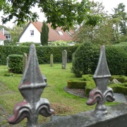 Jüdischer Friedhof Achim Eisenzaun mit Lilien verziert, im Hintergrund alte Grabsteine und umgebende Hecken auf grünem Gelände.