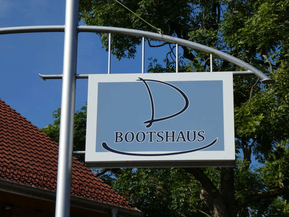 Holzschild mit segelbootförmigem Logo des Bootshaus Hotel-Restaurants vor grünen Bäumen.