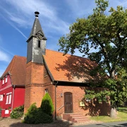 Kapelle Wellie Eine Backsteinkapelle mit Schindelturm und rotem Anbau, umgeben von Bäumen, unter blauem Himmel.