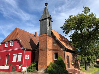 Historische Backsteinkapelle mit Spitzdach und Glockenturm, neben rotem Gebäude, unter blauem Himmel.