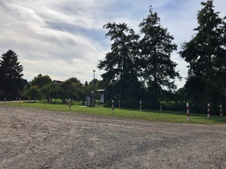 WoMo Steyerberg Wohnmobilstellplatz mit Bäumen, Schotterboden und Pollern in einer grünen Umgebung an einem sonnigen Tag.