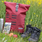 Wanderrast  Wanderausrüstung auf Wiese: roter Rucksack, Thermoskanne, Karte, Apfel, Wanderschuhe vor Rapsfeld.