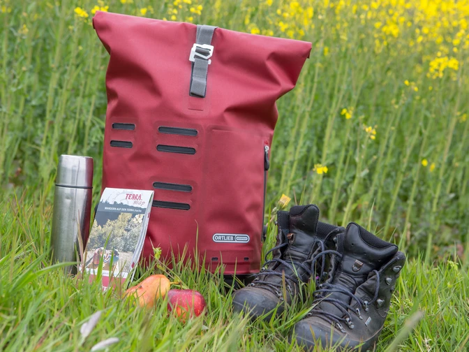 Wanderrast  Wanderausrüstung auf Wiese: roter Rucksack, Thermoskanne, Karte, Apfel, Wanderschuhe vor Rapsfeld.