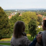 Blick von der Himmelsterrasse Zwei Personen genießen die Aussicht auf eine grüne Parklandschaft mit Kirchturm in der Ferne.