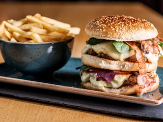 Burger with fries Doppelter Cheeseburger mit Speck, Salat und Käse auf einem Teller, daneben eine Schale mit Pommes frites, auf einem Holztisch serviert.