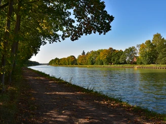 TERRA.track Penter Egge Grüner Weg entlang eines breiten Kanals, gesäumt von Herbstbäumen und blauem Himmel.