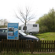 Wohnmobilstellplatz am Dino-Park Stellplatz mit Schild für Wohnmobile und Pkw, umgeben von Bäumen und Wiese mit Löwenzahn.