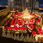 Christmas market at Cologne Cathedral Ein festlich beleuchteter Weihnachtsmarkt mit zahlreichen roten Verkaufshütten unter dem majestätischen Kölner Dom bei Nacht. Der Markt ist dicht besucht, die Atmosphäre ist lebendig und fröhlich. Eine große, strahlend geschmückte Tanne dominiert die Szene.A festively illuminated Christmas market with numerous red sales huts under the majestic Cologne Cathedral at night. The market is crowded, the atmosphere is lively and cheerful. A large, brightly decorated fir tree dominates the scene.