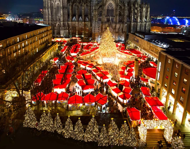 Christmas market at Cologne Cathedral Ein festlich beleuchteter Weihnachtsmarkt mit zahlreichen roten Verkaufshütten unter dem majestätischen Kölner Dom bei Nacht. Der Markt ist dicht besucht, die Atmosphäre ist lebendig und fröhlich. Eine große, strahlend geschmückte Tanne dominiert die Szene.A festively illuminated Christmas market with numerous red sales huts under the majestic Cologne Cathedral at night. The market is crowded, the atmosphere is lively and cheerful. A large, brightly decorated fir tree dominates the scene.