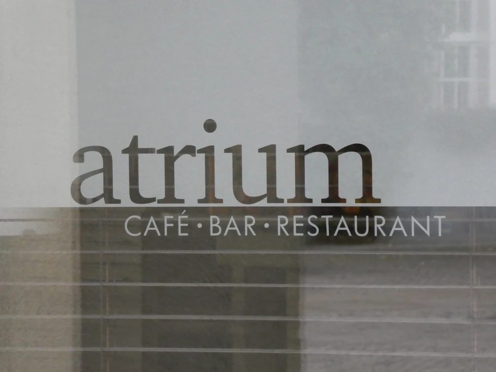 Schriftzug "Atrium Café Bar Restaurant" auf einer Glasoberfläche, dahinter verschwommene Gebäudestrukturen.