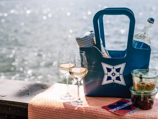 Picknick Zwei Gläser Weißwein, ein gefüllter Picknickkorb und Plastikbehälter auf einem Tuch am Seeufer.