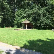 Ein hölzerner Unterstand auf einer Grasfläche, umgeben von Bäumen, mit mehreren Holzbänken.