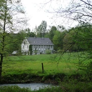 Brucher Kotten von der Itterseite aus Historisches Fachwerkgebäude am Fluss, umgeben von grüner Wiese und Bäumen im Hintergrund.