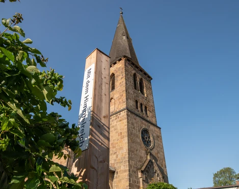 Bibel-Pfahl 41 - Bad Driburg Historische Kirche mit hohem Turm, flankiert von einem modernen Pfahl mit Bibelzitat, klarer Himmel.