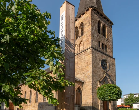 Bibel-Pfahl 41 - Bad Driburg Pfahl mit Bibelvers vor historischer Kirche, umgeben von grünem Rasen und blauem Himmel.