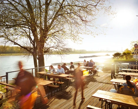 Haus Rheinblick Ott Eine belebte Terrasse am Flussufer des Rheins mit Menschen, die an Tischen speisen und genießen.
