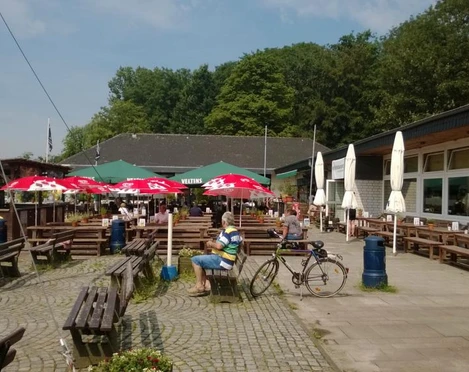 Se(h)-Restaurant Unterbacher See Sonnige Terrasse eines Restaurants am See mit rot-weißen Sonnenschirmen, Holzsitzbänken und Fahrrädern.