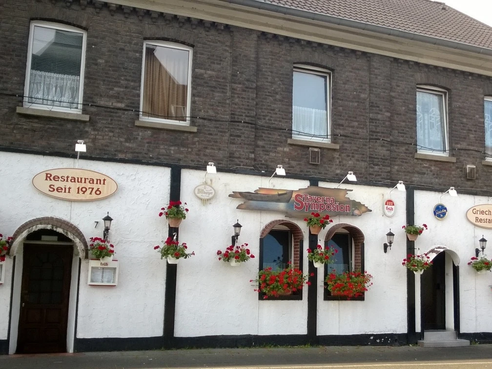 Taverna Symposion Das Bild zeigt ein traditionelles Restaurantgebäude mit weißen Wänden und dekorativen Blumen.